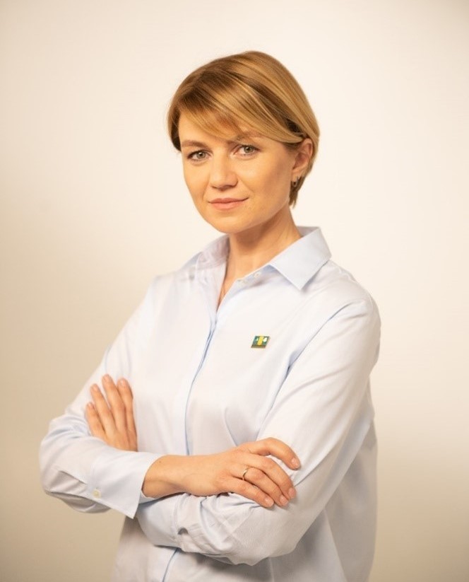 Halyna Scipalska standing with arms crossed in front of a beige photo studio background; determined look on a blond haired woman 